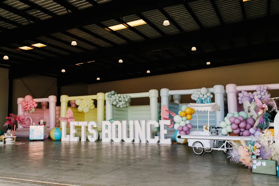 Pastel bounce houses behind LETS BOUNCE marquee letters with balloon garlands and a popsicle cart inside The Executive Hangar