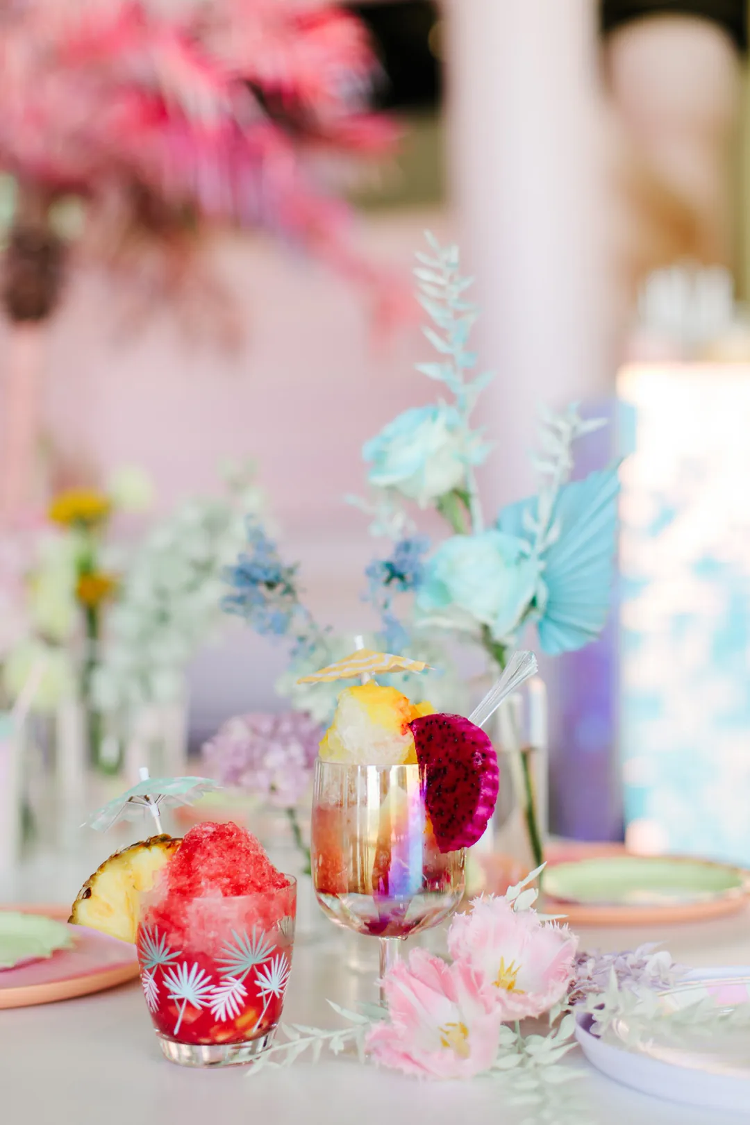 Two colorful shaved ice cups garnished with pineapple, dragon fruit and paper umbrellas on a table with pastel blue florals