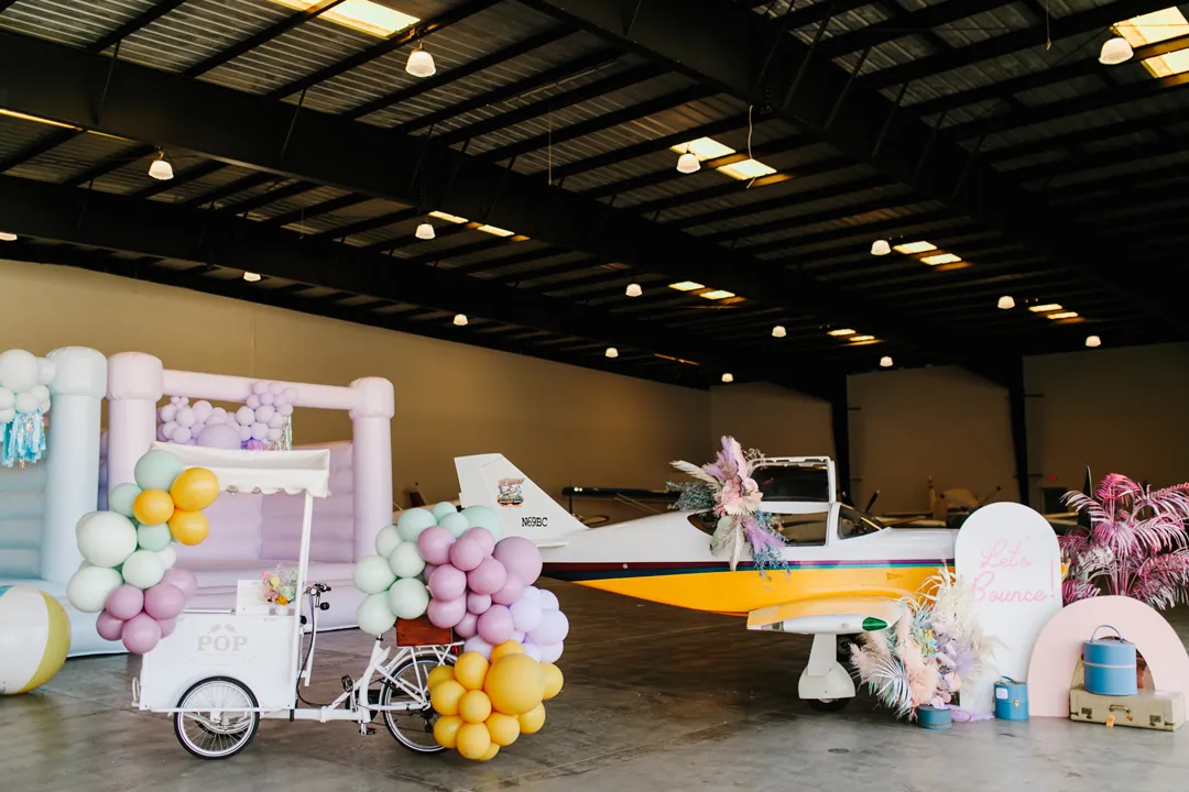 POP popsicle tricycle with balloon garlands beside a floral-decorated plane and pastel bounce house in The Executive Hangar
