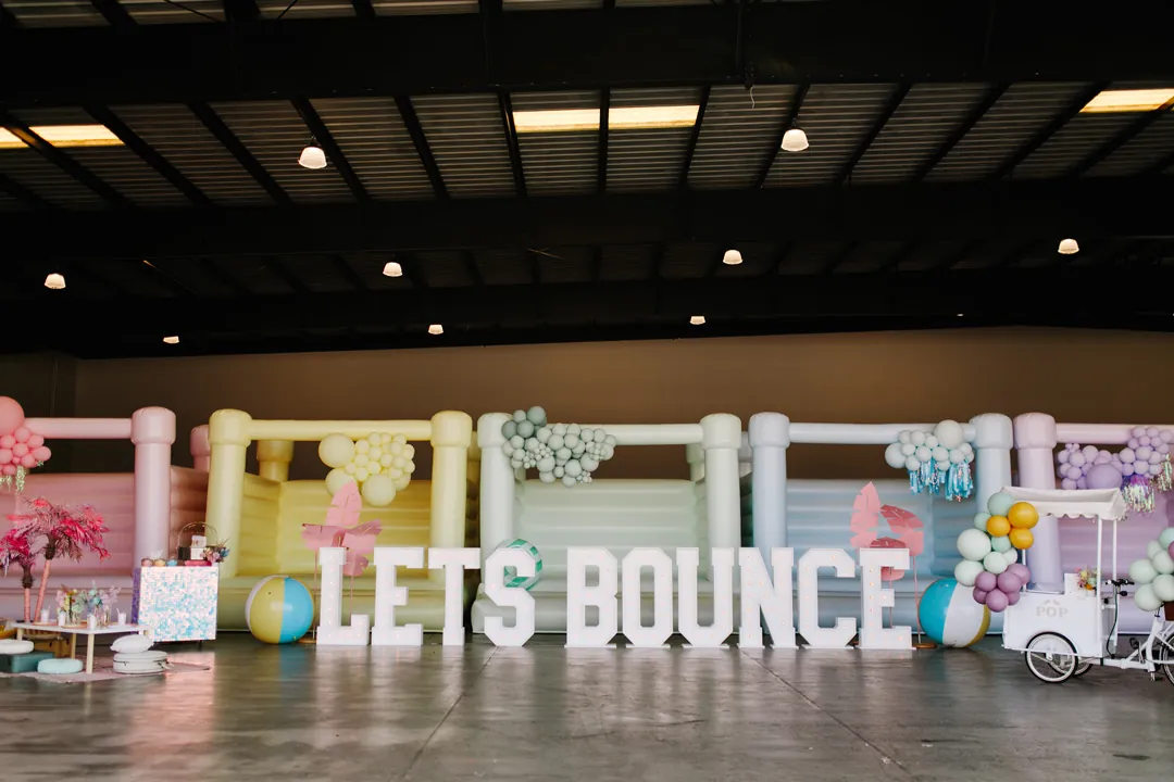 Row of five pastel bounce houses with balloon garlands behind LETS BOUNCE marquee letters and beach balls at the hangar