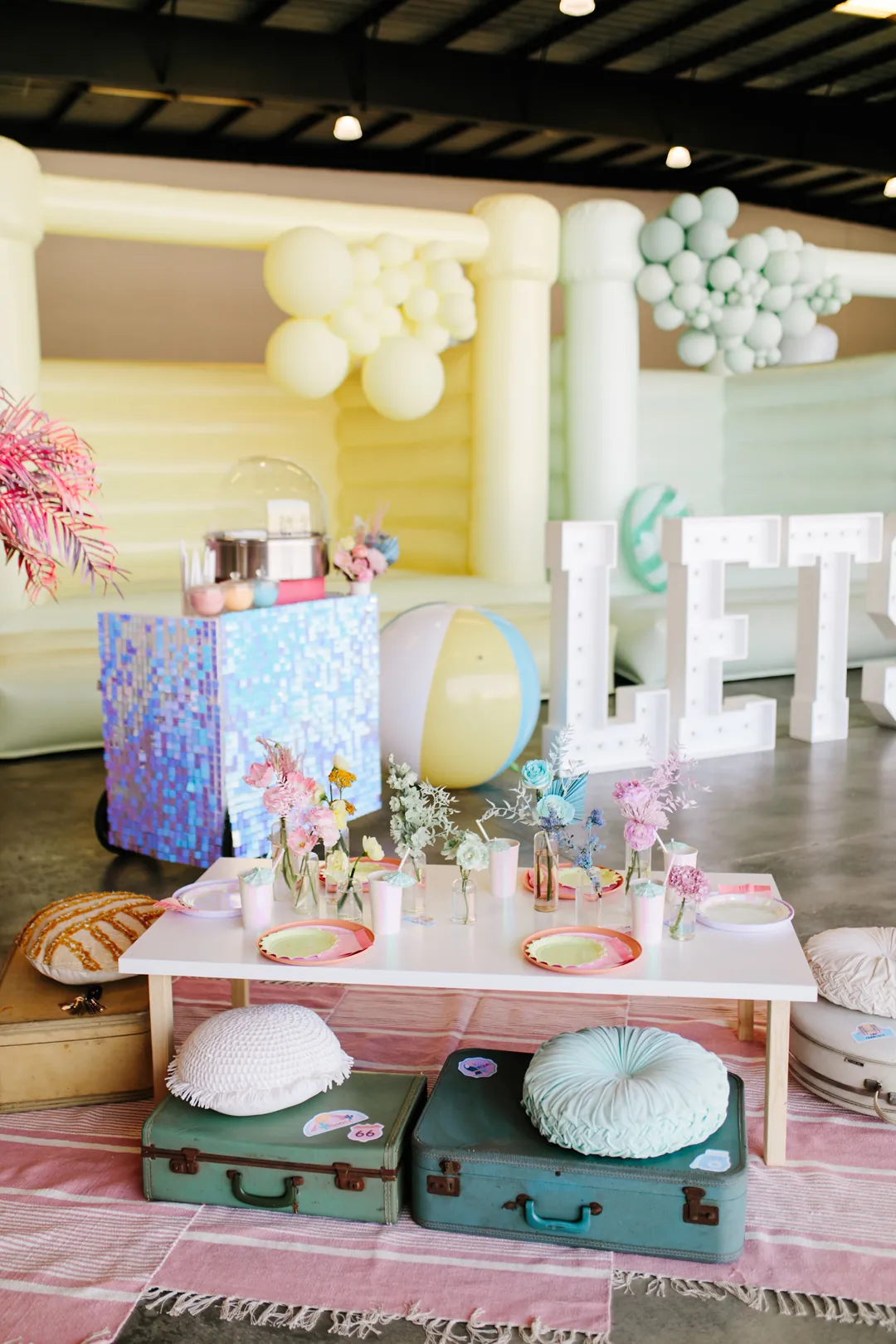 Kids table with pastel plates, rainbow florals, a beach ball, cotton candy cart and LETS marquee letters by a yellow jumper