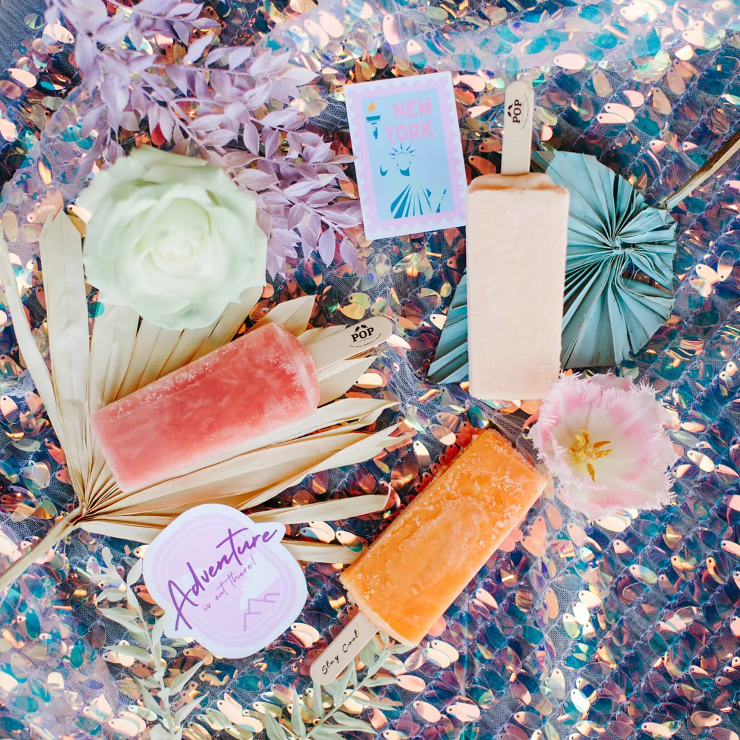 Flat lay of three fruit popsicles, a white rose, dried palm leaves and travel stickers on iridescent sequin fabric