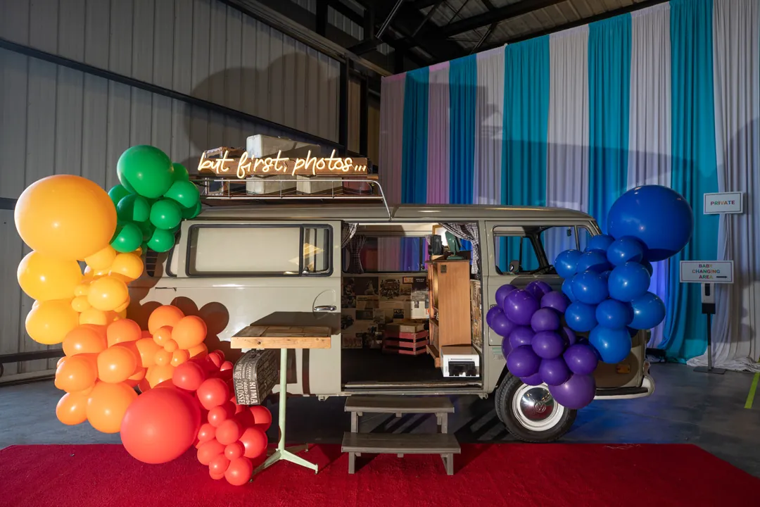 Vintage VW bus photo booth with a but first photos neon sign and rainbow balloon garlands inside The Executive Hangar