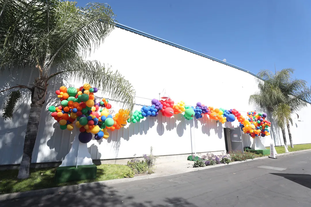 Rainbow balloon garland stretched along the white exterior wall of The Executive Hangar, framed by palm trees