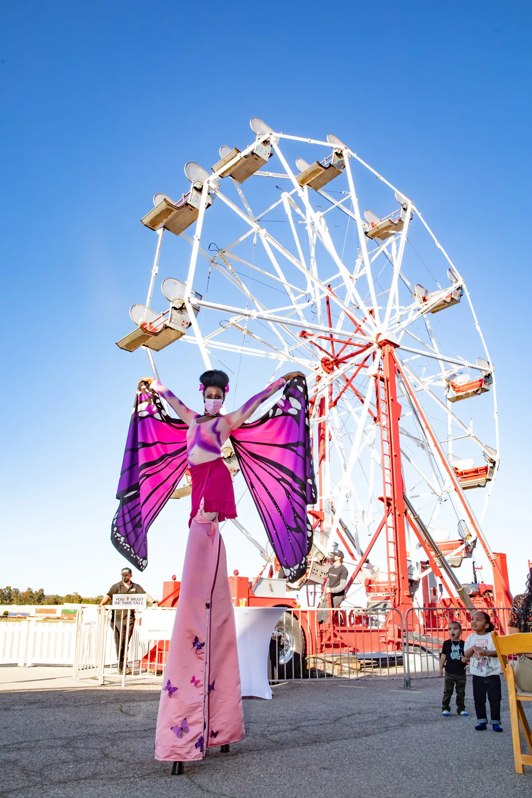Stilt walker in pink butterfly costume performing in front of the ferris wheel on the tarmac at The Executive Hangar