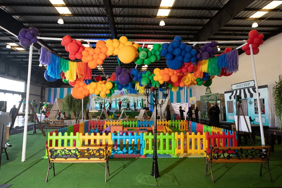 Rainbow balloon canopy over a fenced toddler play zone with benches and a Sweet Shop facade inside The Executive Hangar