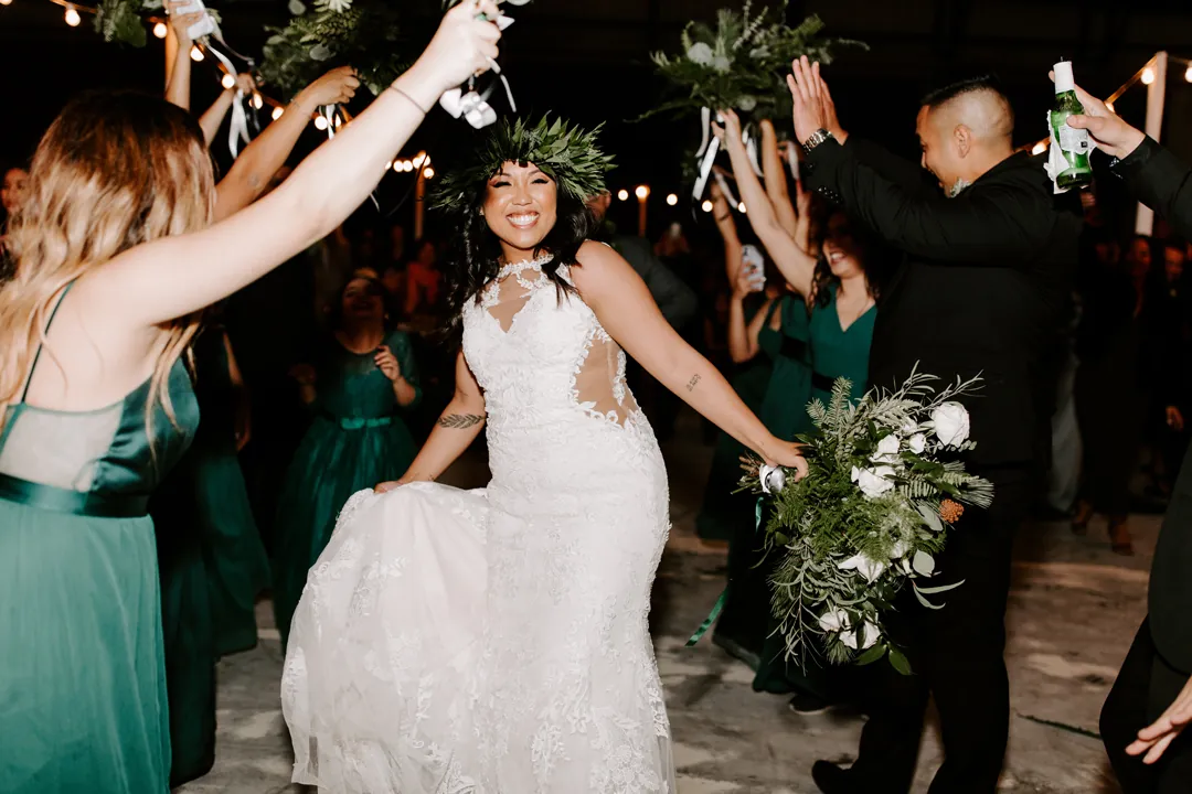 Roni grins as bridesmaids in emerald green raise bouquets over her during the grand entrance into the hangar