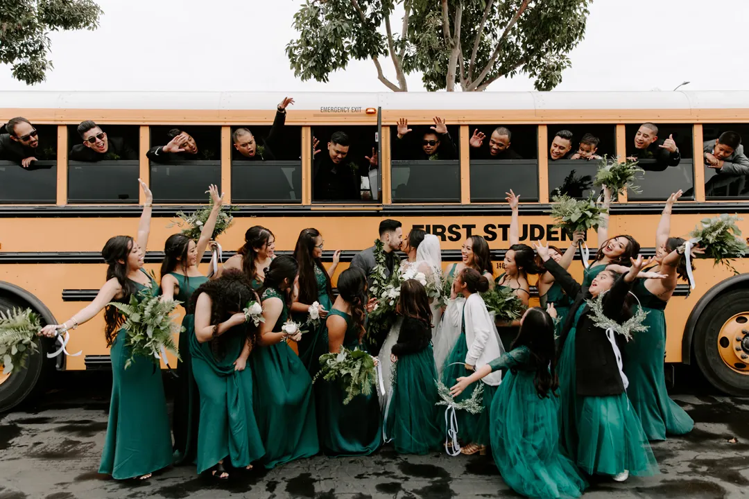 Roni and James kiss in front of a yellow school bus as bridesmaids in emerald green cheer at The Executive Hangar