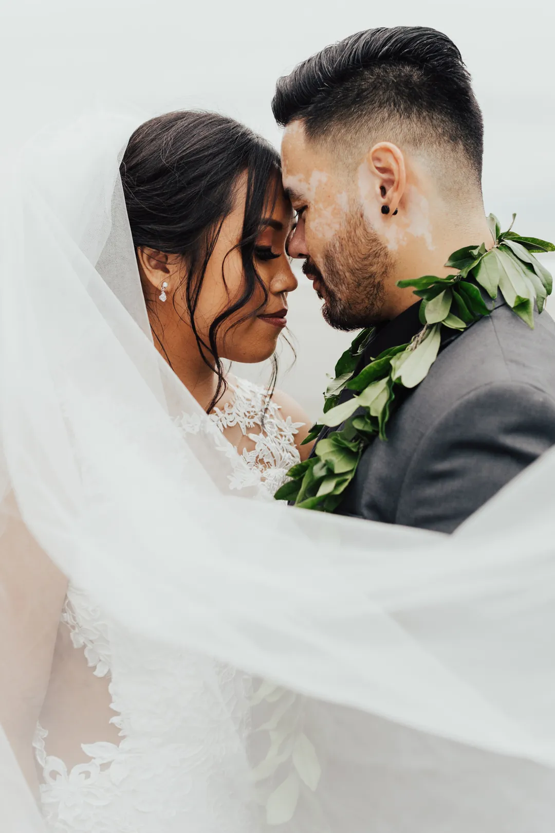 Roni and James touch foreheads under a lace veil, the groom wearing a green maile lei over a grey suit