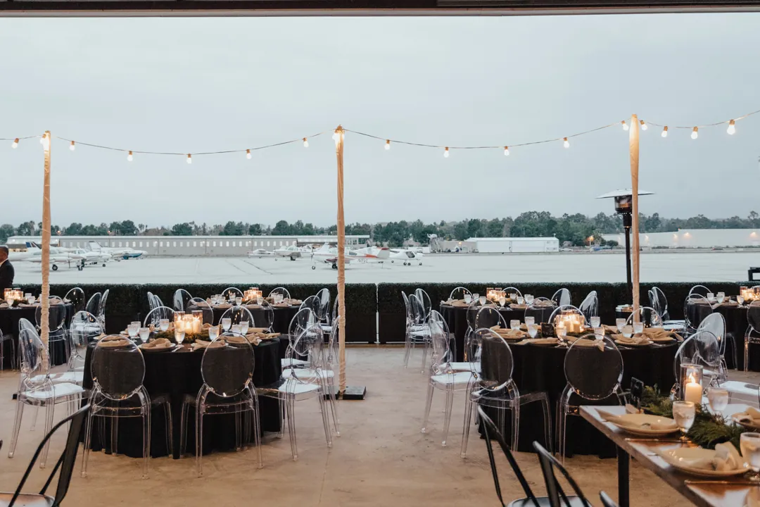 Black-draped round tables and ghost chairs under string lights facing the tarmac and parked planes at The Executive Hangar
