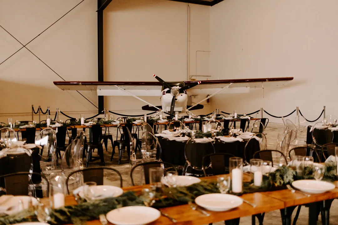 Wedding reception tables with evergreen garland set in front of a small white airplane inside The Executive Hangar