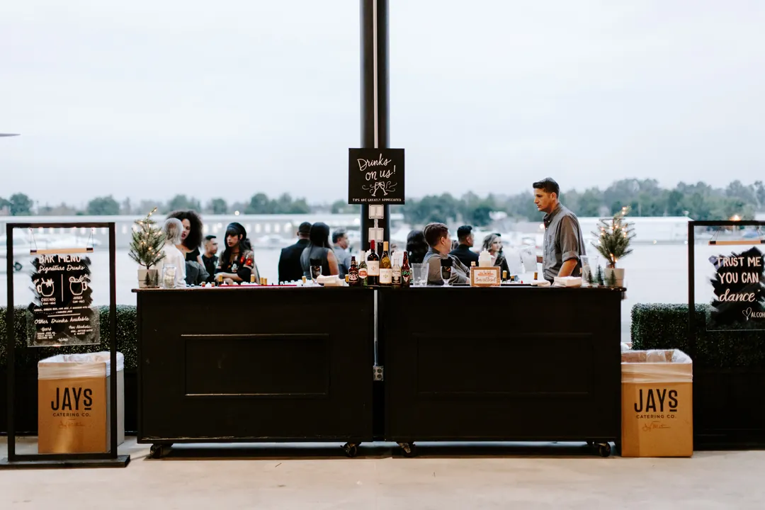 Jay's Catering bartender at a black rolling bar with acrylic menu signs and mini Christmas trees at The Executive Hangar