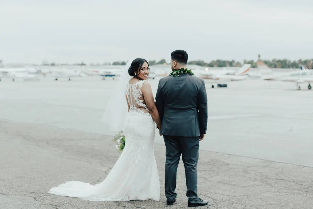 Roni in a lace gown and James in a grey suit hold hands on the tarmac with planes lined up behind them