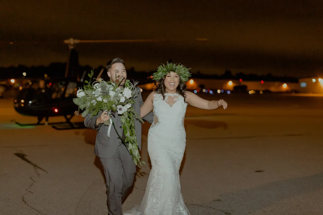 Roni in a haku lei and James carrying her bouquet walk from the helicopter across the tarmac at night