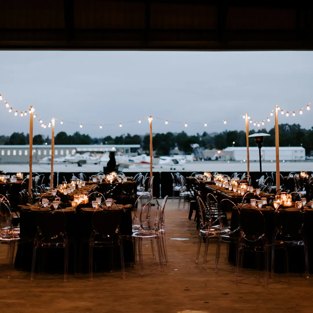 Candlelit reception tables and ghost chairs under string lights with the hangar door open to the tarmac at dusk