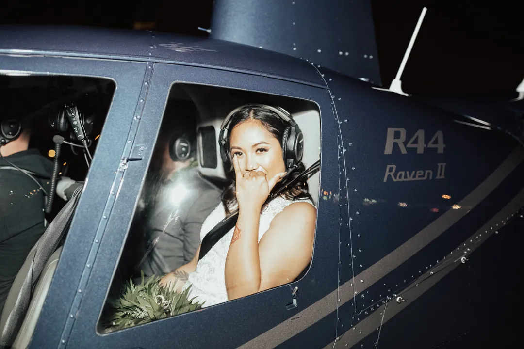 Roni wearing a headset smiles and flashes a shaka from the window of an R44 helicopter at night