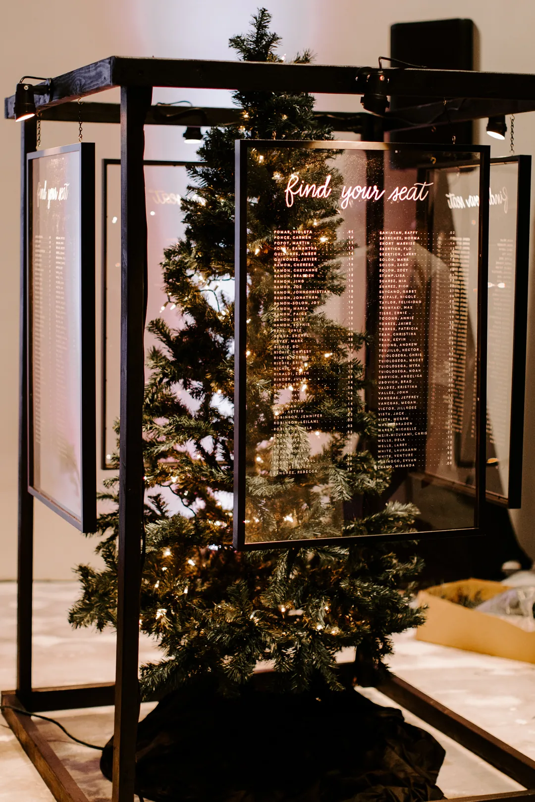 Lit Christmas tree inside a black frame with acrylic Find Your Seat panels serving as the wedding seating chart