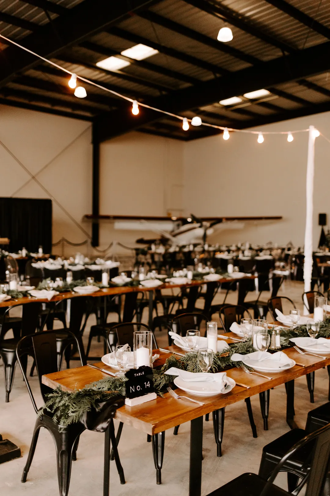 Long farmhouse tables with black chairs, evergreen garland and table number cards under bistro lights in the hangar