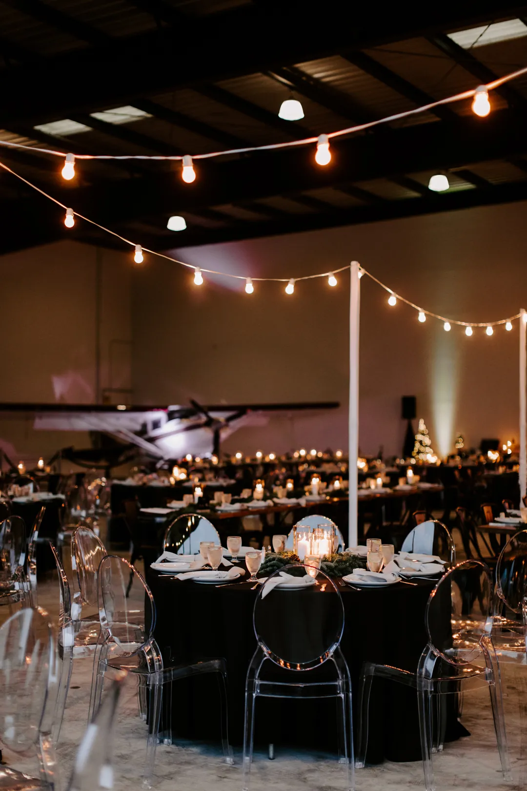 Round black-draped table with ghost chairs and candles under string lights, with an airplane and Christmas tree behind