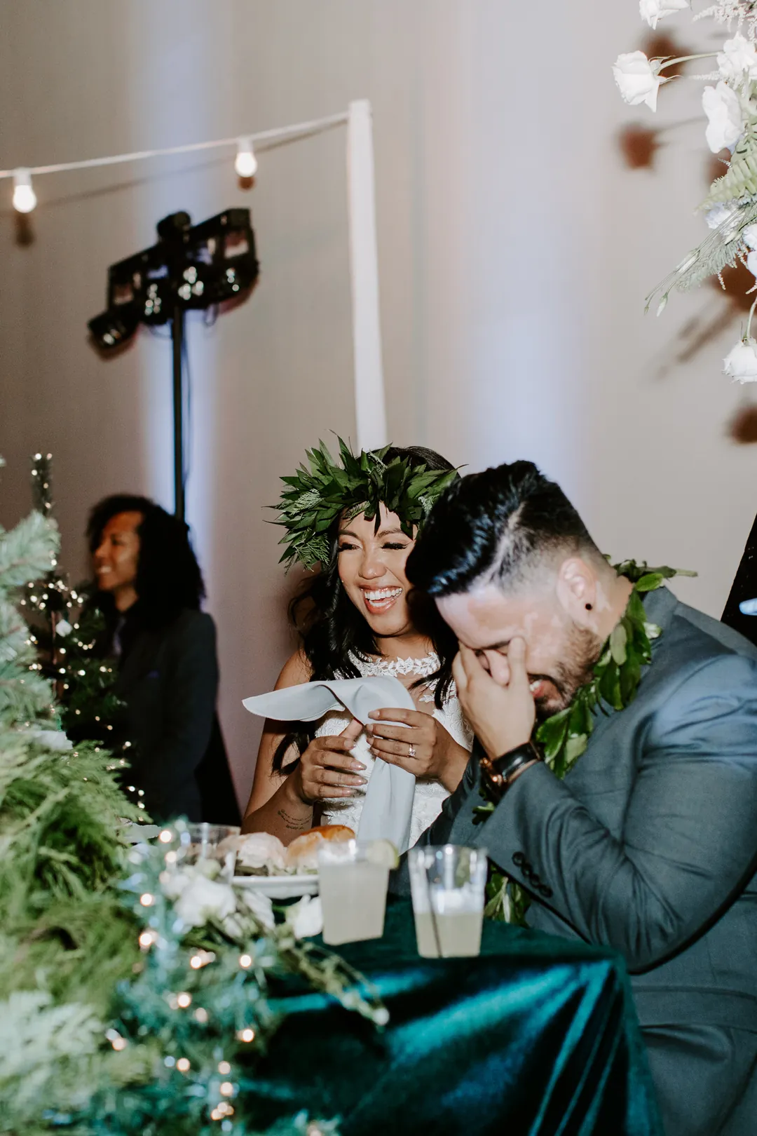 Roni laughs and James covers his face at the velvet-draped sweetheart table during wedding toasts