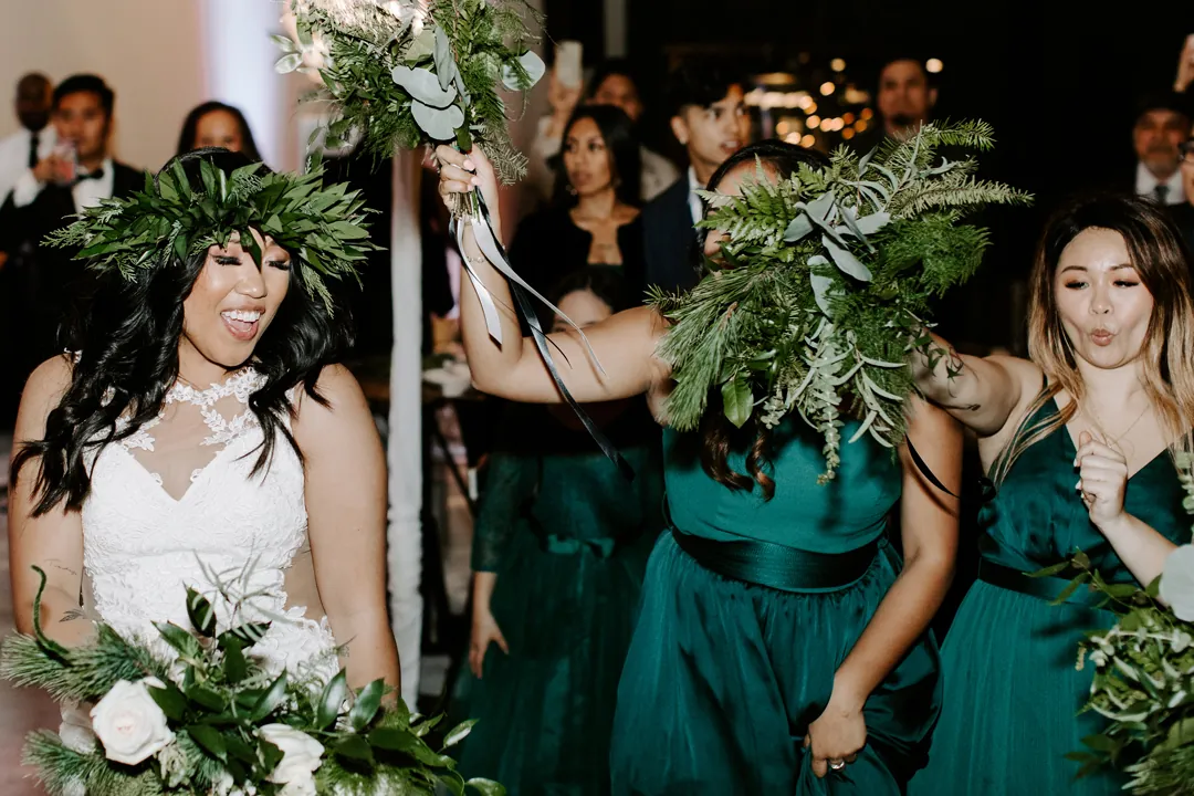 Roni laughs beside bridesmaids in emerald gowns waving greenery bouquets during the grand entrance