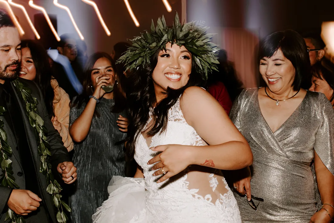 Roni in a haku lei laughs on the hangar dance floor surrounded by smiling guests and a lei-wearing groomsman
