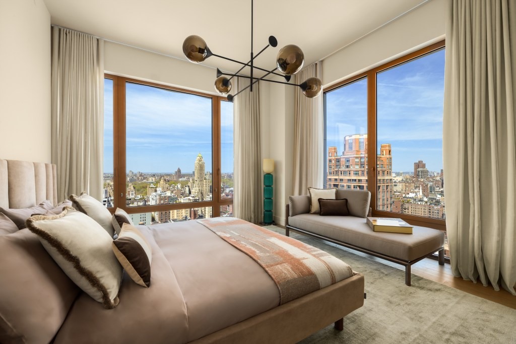 A spacious bedroom with a king-sized bed, chaise lounge, and floor-to-ceiling windows offering a panoramic city view.