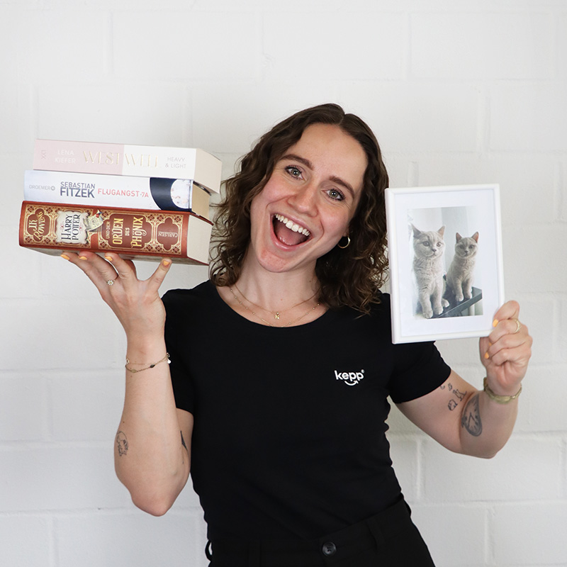 Porträt von Natalie Hornoff mit einem Bücherstapel in der einen und einem Bild von zwei Katzen in der anderen Hand
