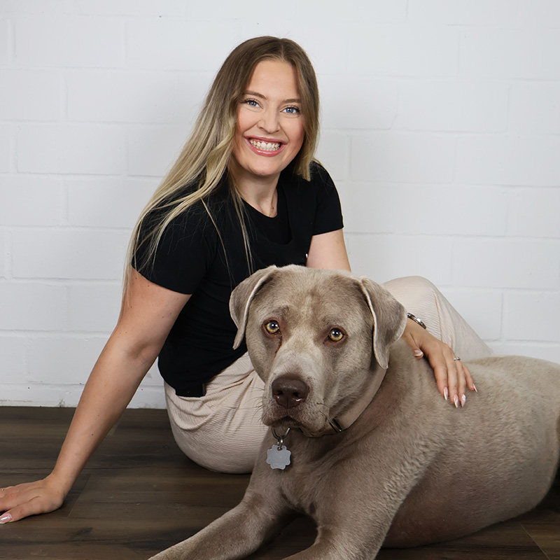 Porträt von Hanna Lalla auf dem sie lächelt, auf dem Boden neben ihrem Hund sitzt und ihn streichelt