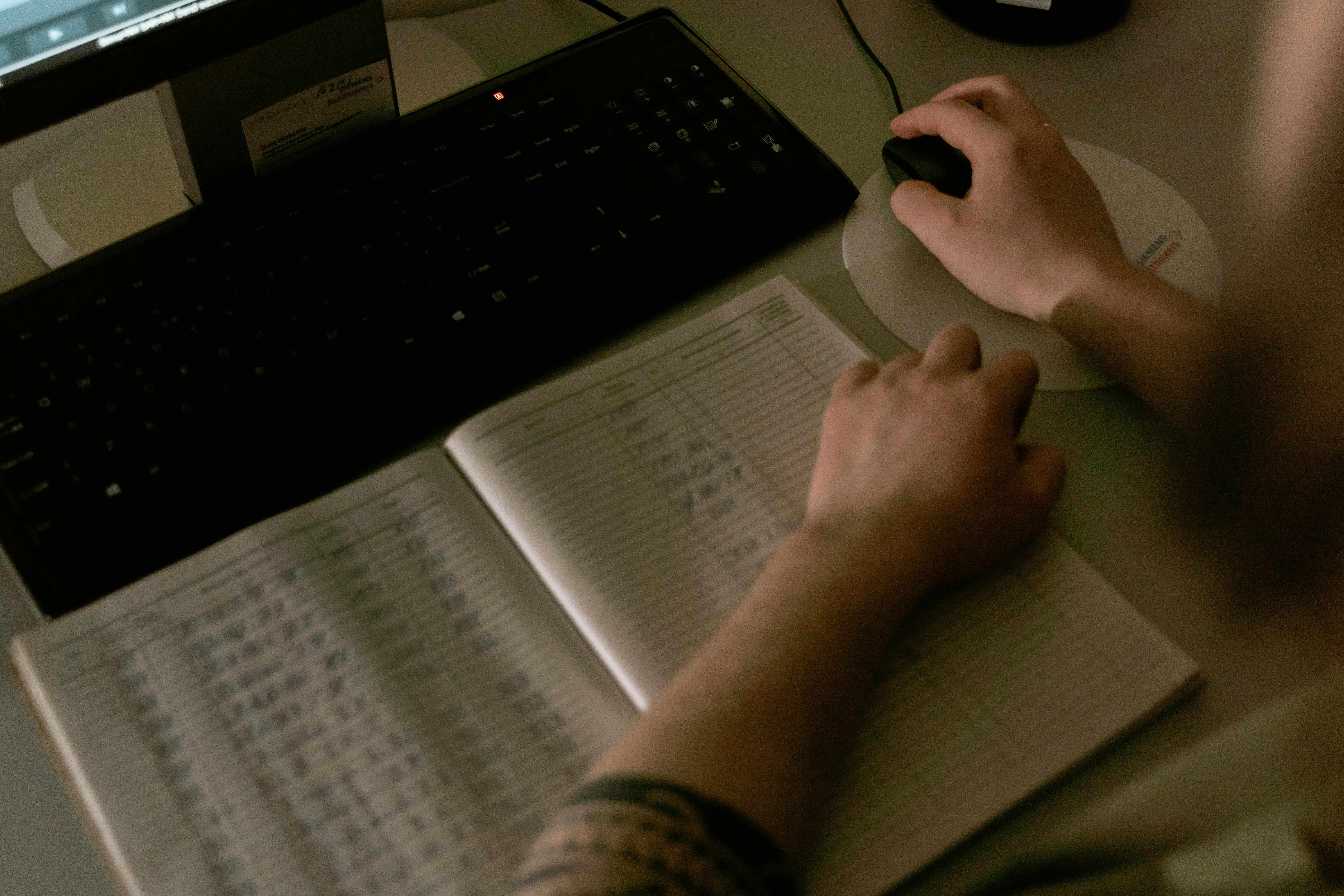 Hands working at a desk with a keyboard, mouse, and spreadsheet, representing manual product data management work.