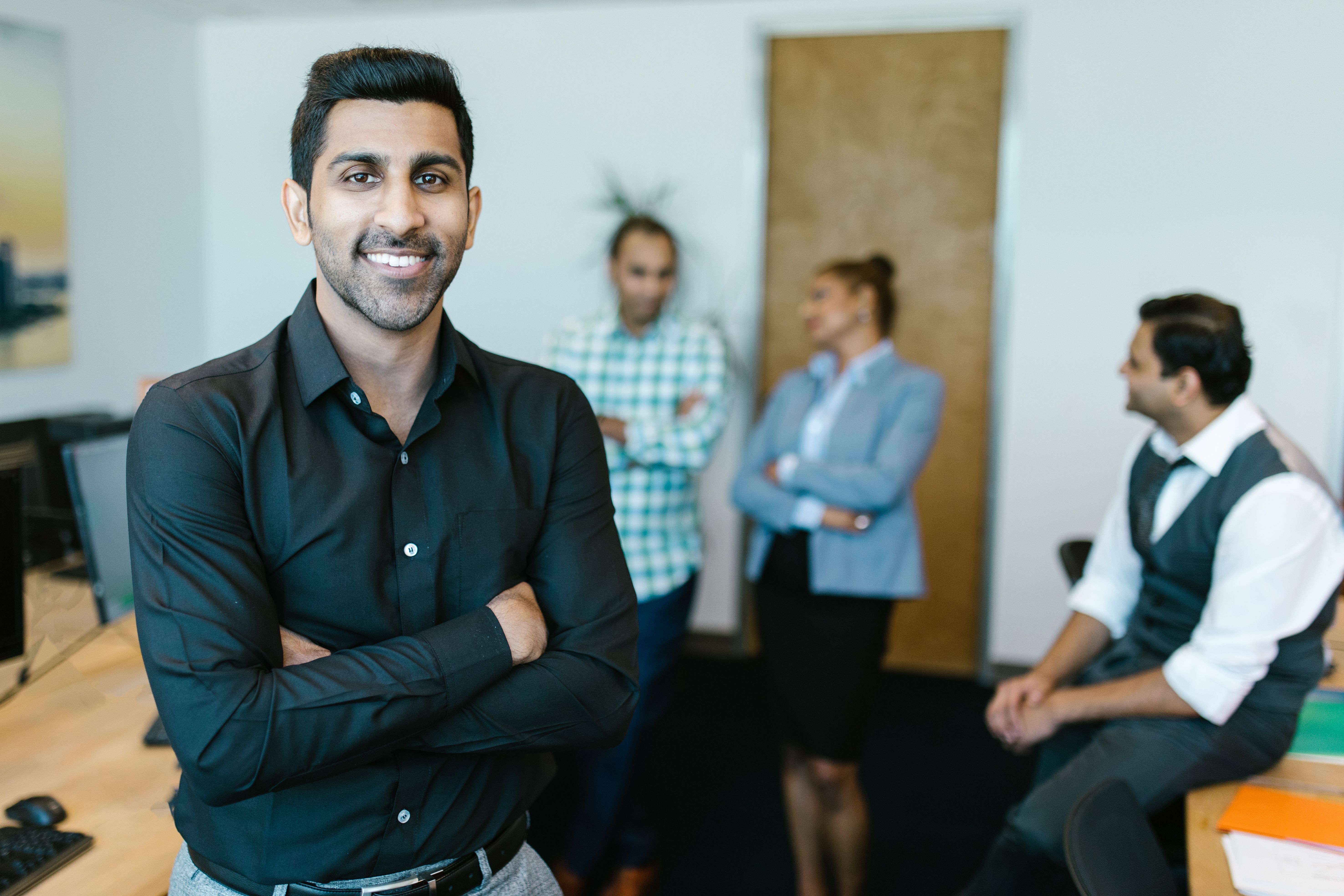 Man with arms crossed smiling at the camera in an office, with colleagues talking in the background.