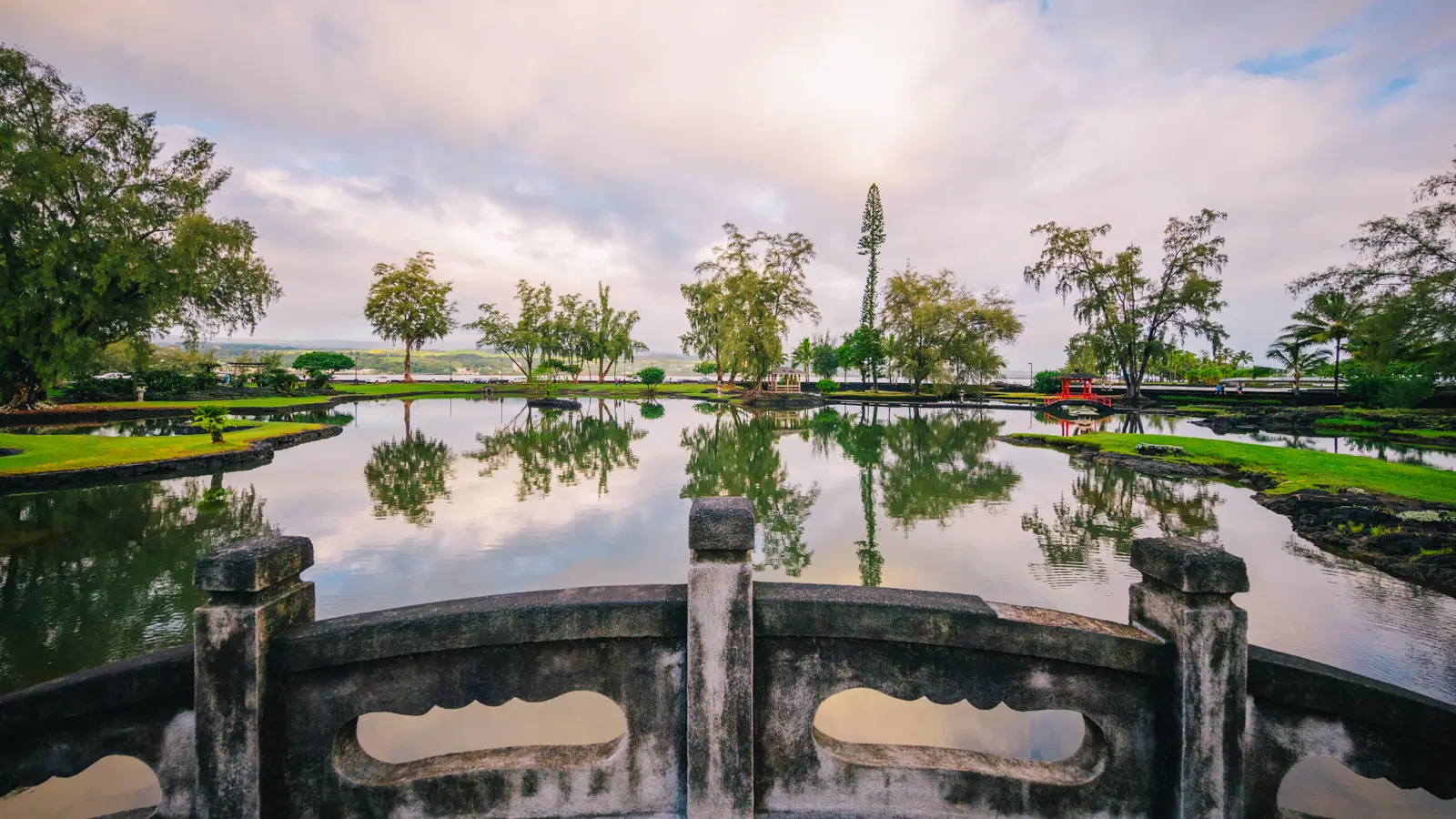 Scenic view of a serene park featuring a reflective pond surrounded by lush trees and a pedestrian bridge.