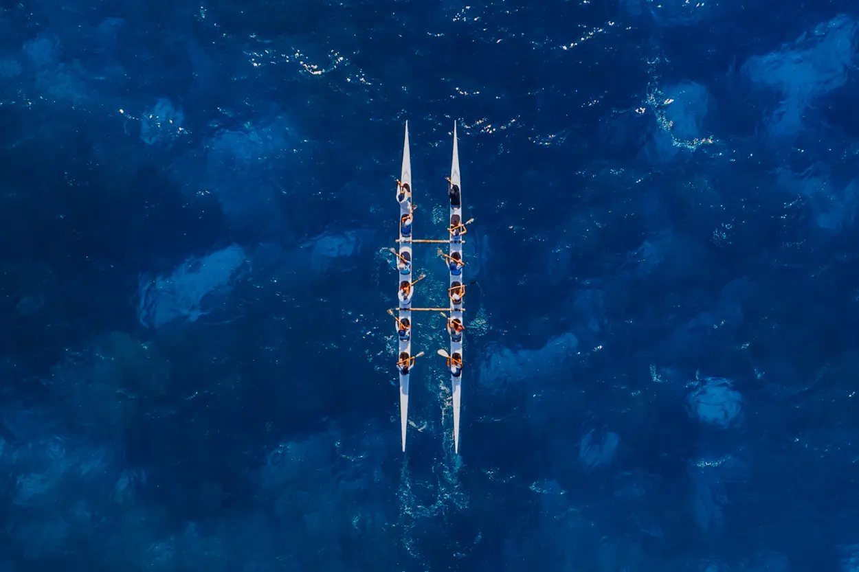 Aerial view of two outrigger canoes with teams paddling in sync over the deep blue ocean waters.