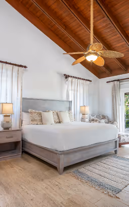 Bright bedroom with wooden ceiling, ceiling fan, grey bed with white bedding, bedside tables with lamps, and large windows with curtains.