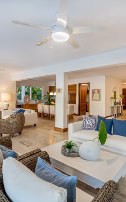 Bright, spacious living room with white sofas, blue cushions, wicker chairs, and a ceiling fan with a light fixture.