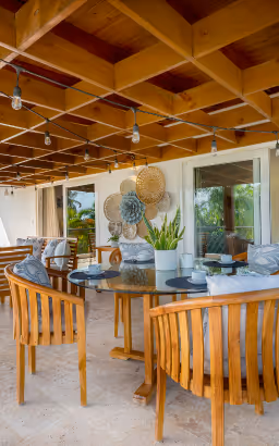 Outdoor dining area with wooden chairs, a round glass table, potted plants, and decorative woven wall hangings under a wooden lattice ceiling.