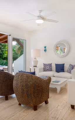 Bright living room with white sofa, blue and patterned cushions, two woven armchairs, white coffee table, and large round mirror on wall.