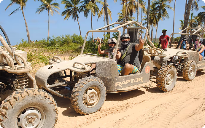 Two people wearing helmets and masks sitting in a muddy off-road buggy on a sandy trail with palm trees in the background.