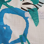 Abstract textile art with shapes in blue, green, and black on a beige fabric background, including a small white patch with handwritten text.