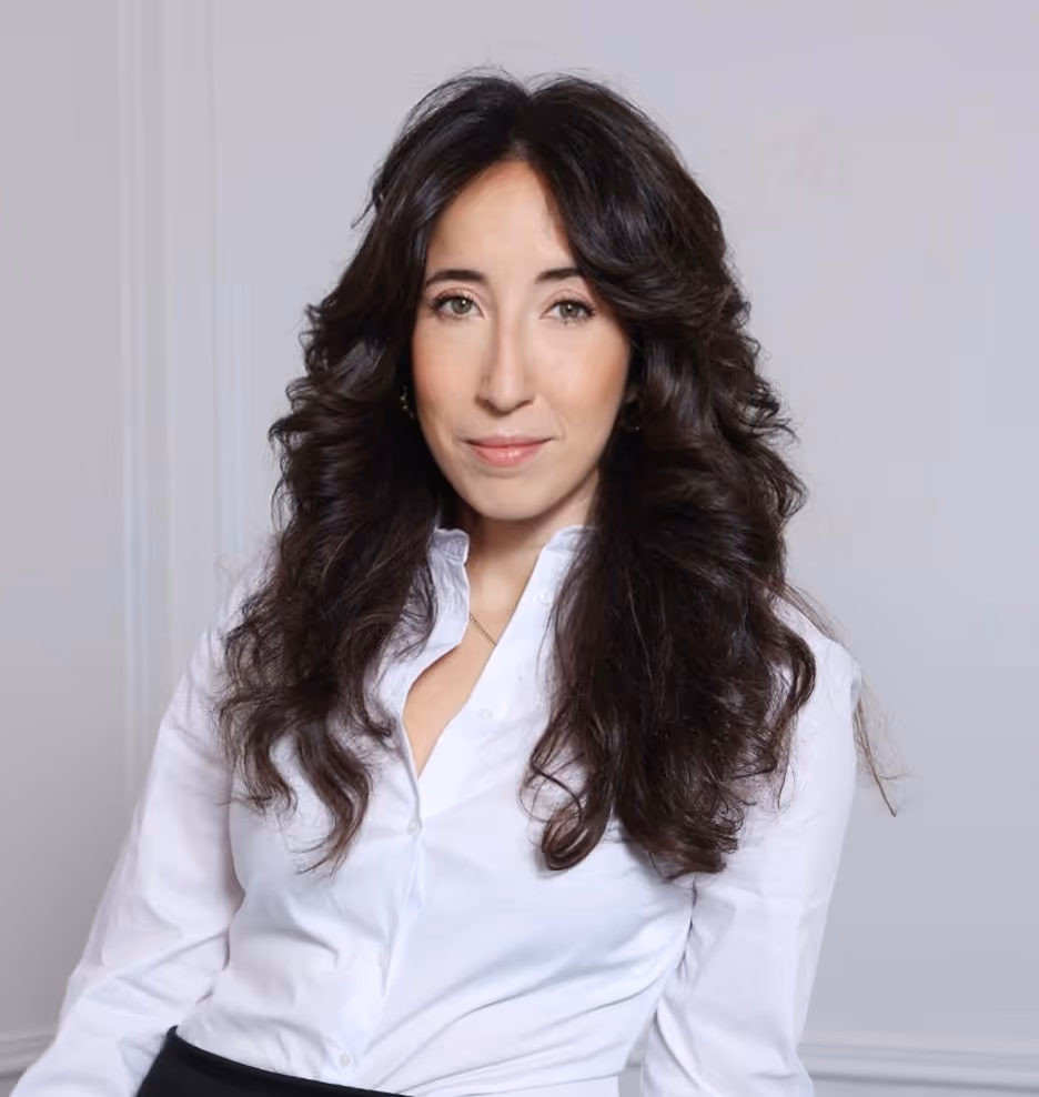 Woman with long curly dark hair wearing a white blouse against a plain gray background.
