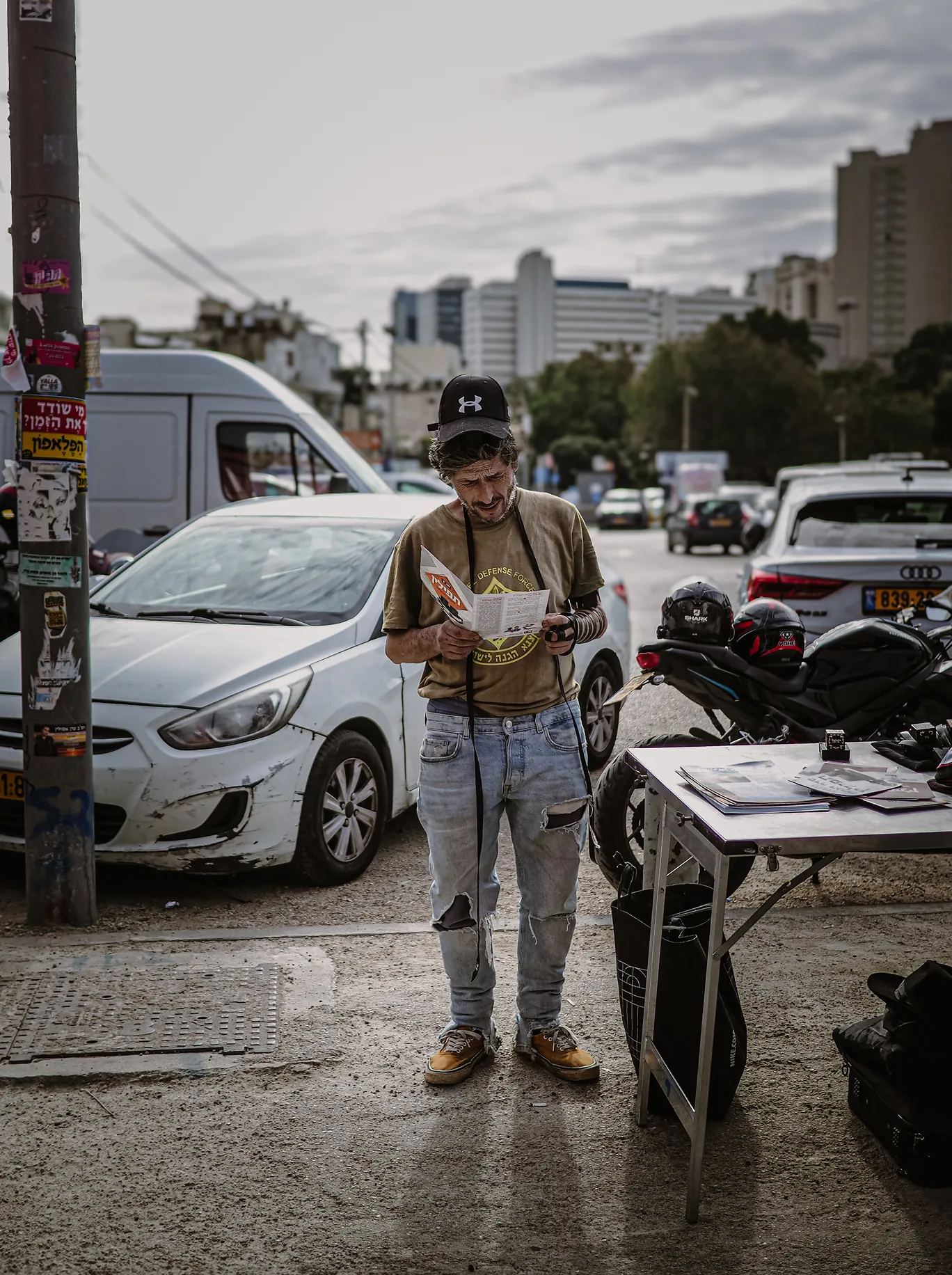 Man wearing a cap and tefillin reading a leaflet on a city street near parked vehicles and a table.