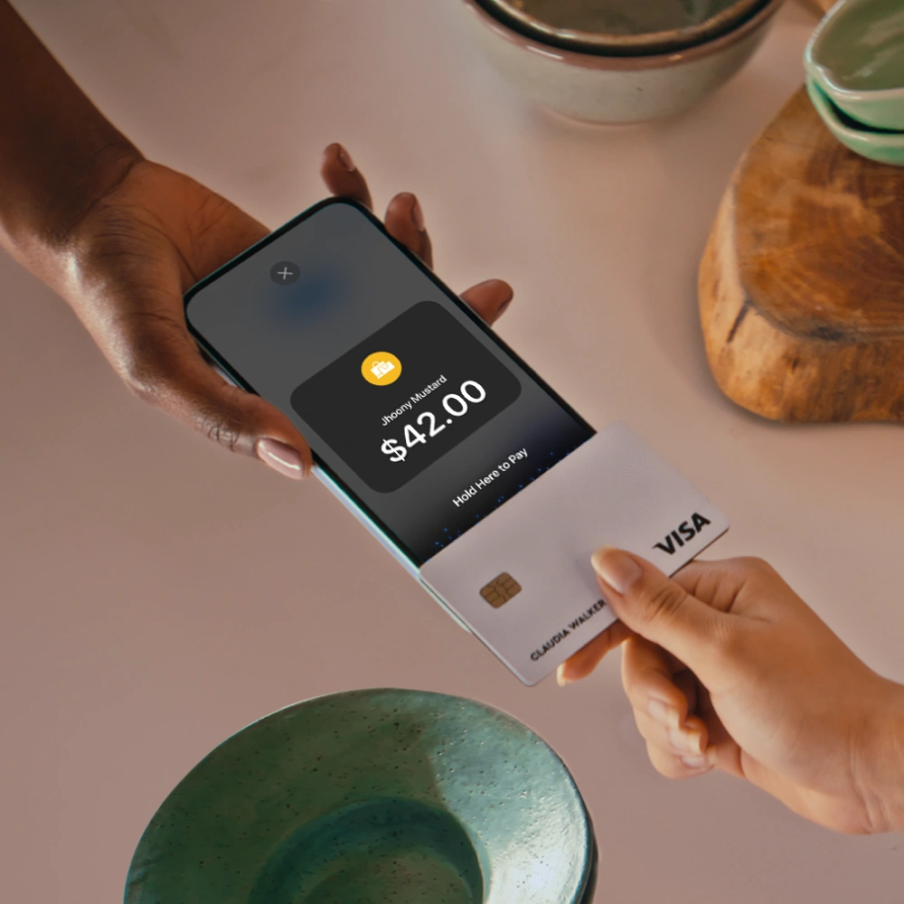 A person holding a smartphone running the JIM app, displaying a payment confirmation screen for $42.00, while another person taps a Visa card on the phone's card reader to complete the transaction.