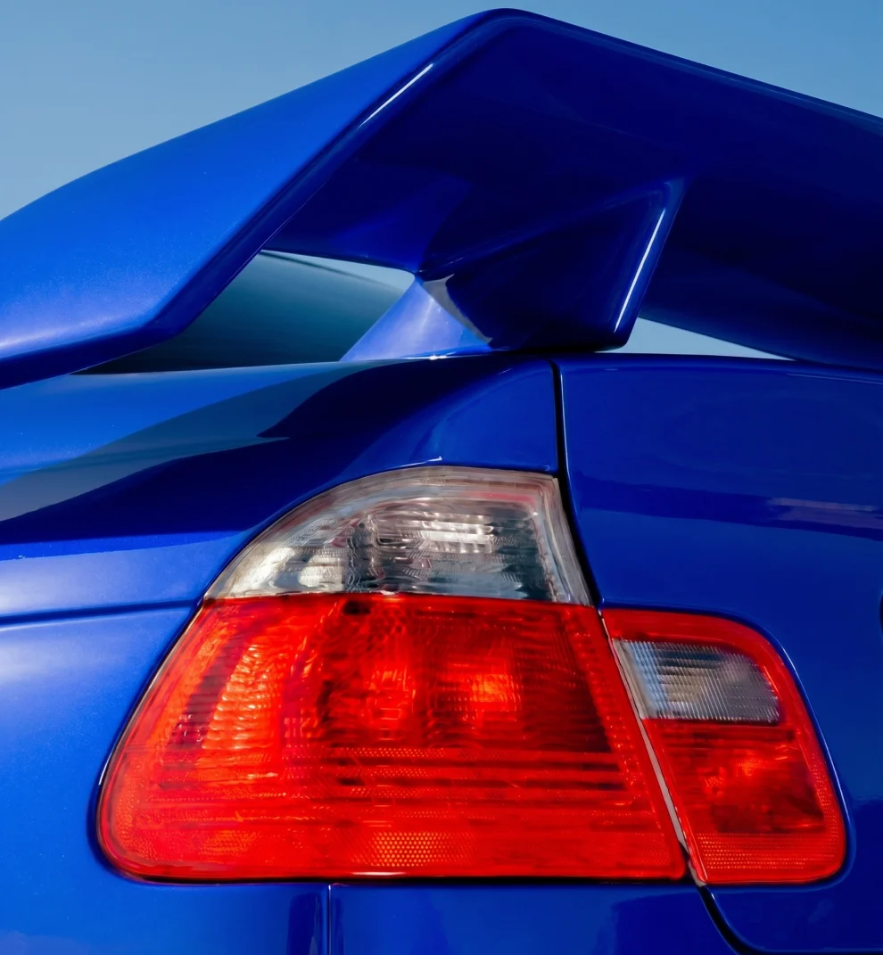 Close-up of a blue car's rear showing red tail light and large spoiler against a clear sky.