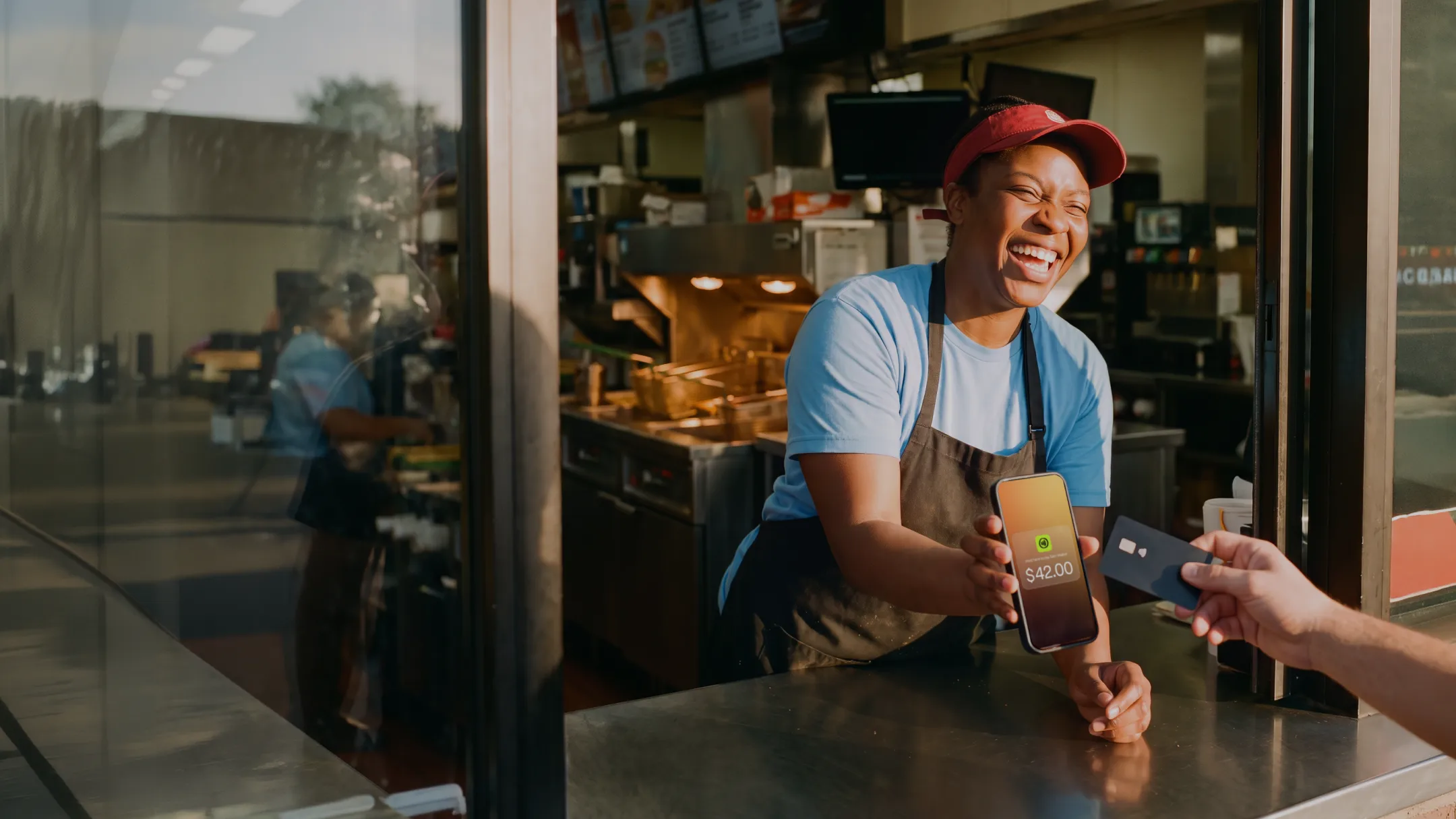 Smiling fast food worker holding phone showing $42.00 while customer pays with a credit card.