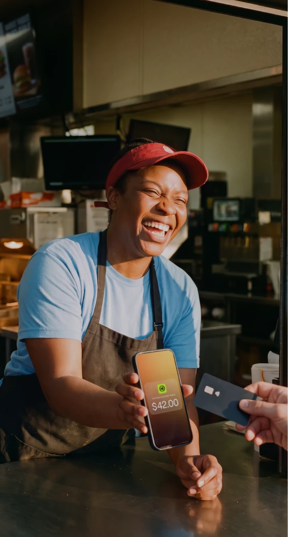 Smiling worker in a red visor shows a phone screen with $42 payment while holding a card at a counter.