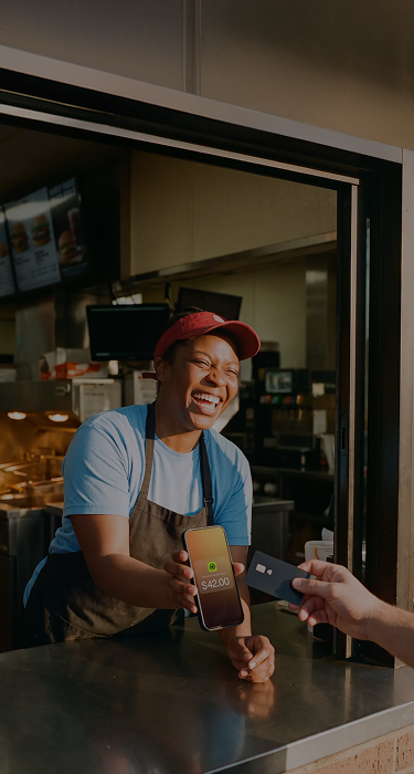 Smiling food service worker holding a phone showing $42 payment as customer hands a credit card.