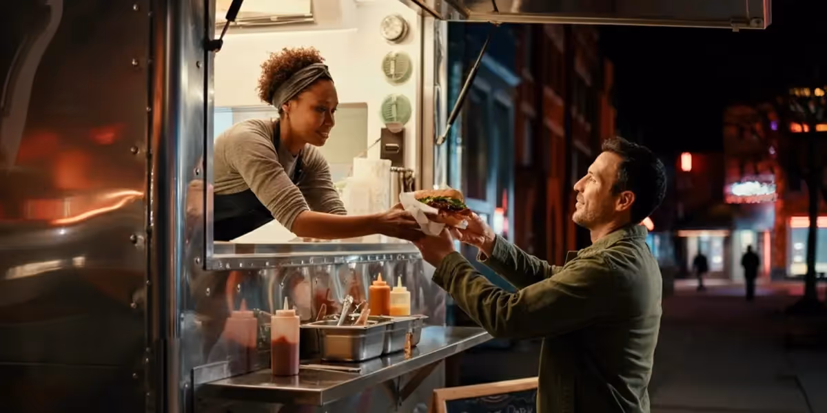 How to start a food truck business: vendor handing burger through window to customer at night.