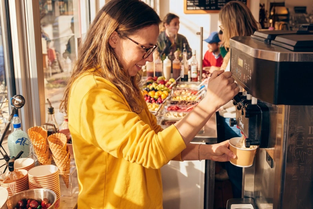 Woman in yellow sweater serving herself from a soft-serve machine in a bright food shop.
