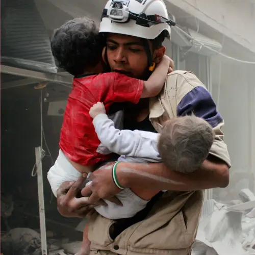 Rescue workers evacuating children from a disaster-stricken area amid rubble and dust.