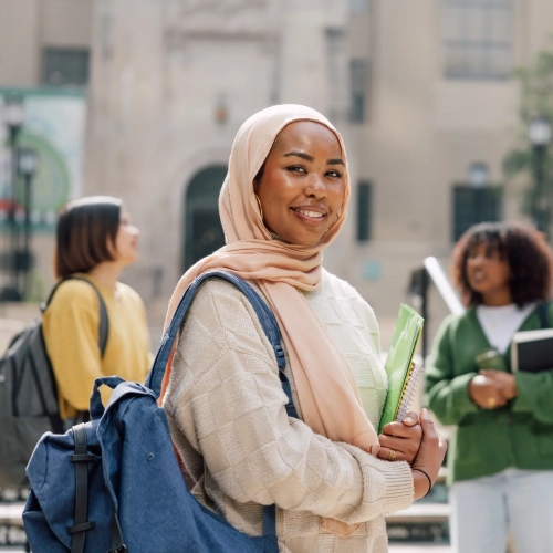 University student carrying books and a backpack on campus, smiling while standing outdoors with fellow students in the background.